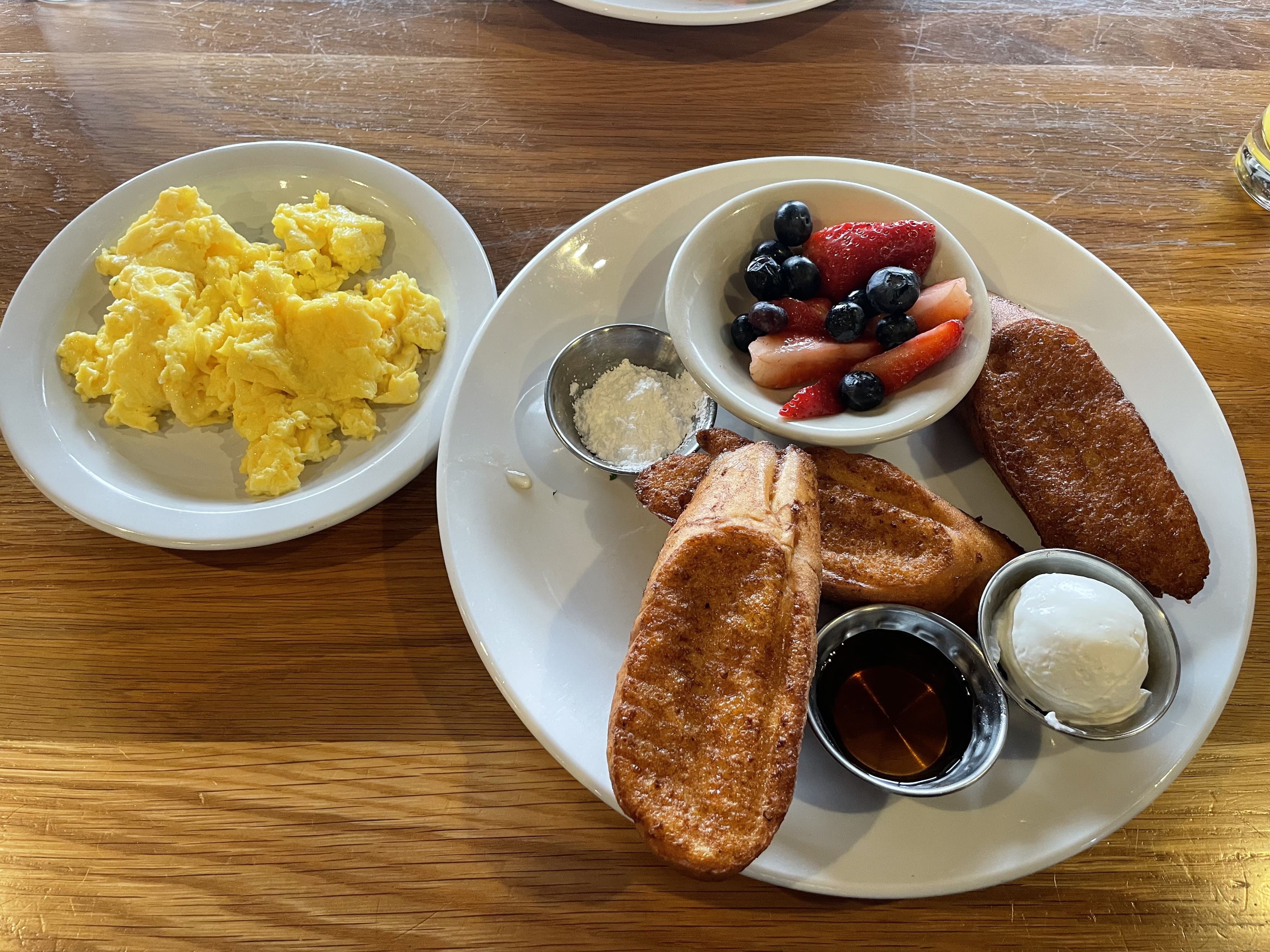 a breakfast plate of french toast, fresh strawberries and blueberries, powdered sugar, syrup, butter, and a side of eggs